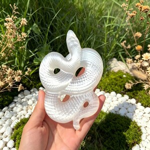 以下が含まれることがあります： 半透明の白いセレナイトを彫り込んだ、とぐろを巻いたヘビの置物。表面は滑らかに磨かれ、体には鱗の模様が刻まれています。精巧な頭部と曲線を描く尾が特徴で、真珠のような光沢を放っています。