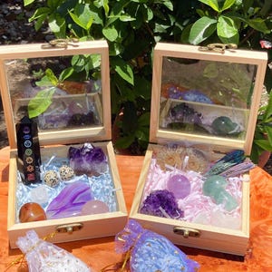 Op de afbeelding: Twee lichtgekleurde houten kisten met glazen deksels, elk gevuld met diverse kristallen en stenen. De linkerdoos bevat een zwarte chakratoren, amethistcluster, agaatplak en kleine eieren. De rechterdoos bevat ammonietfossielen, amethist en een groene paddenstoelvormige steen.