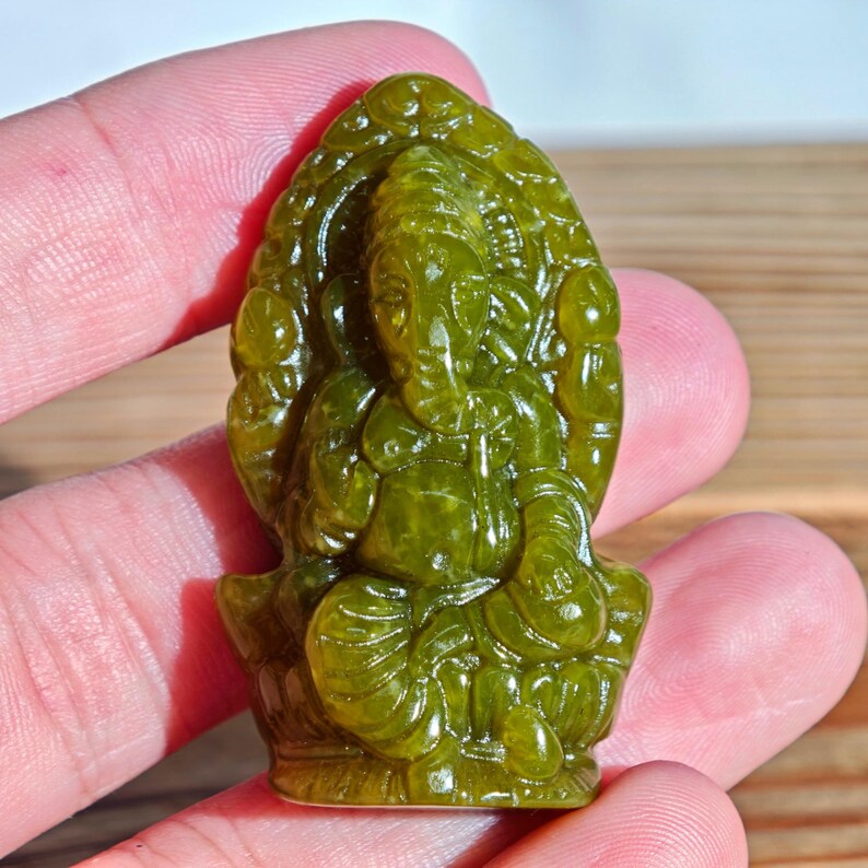 Puede incluir: Una talla de jade verde del dios hind&uacute; Ganesha, sentado en posici&oacute;n de loto. La escultura est&aacute; detallada con rasgos faciales visibles, una gran barriga y m&uacute;ltiples brazos. La talla se sostiene en una mano, mostrando su tama&ntilde;o y dise&ntilde;o intrincado.