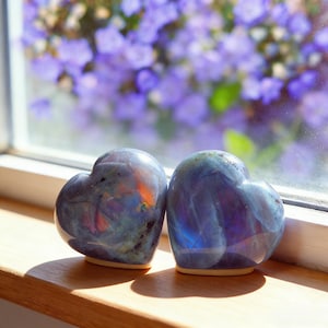 Puede incluir: Dos piedras de labradorita en forma de corazón con tonos iridiscentes azules, morados y anaranjados. Las piedras pulidas están sobre un alféizar de madera, con un fondo borroso de flores moradas.