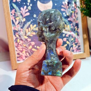 Puede incluir: Una escultura de busto de labradorita tallada. La escultura es una cabeza humana estilizada con una apariencia detallada de calavera. La piedra tiene una mezcla de tonos grises, azules y verdes. El busto est&aacute; montado sobre una base rectangular.