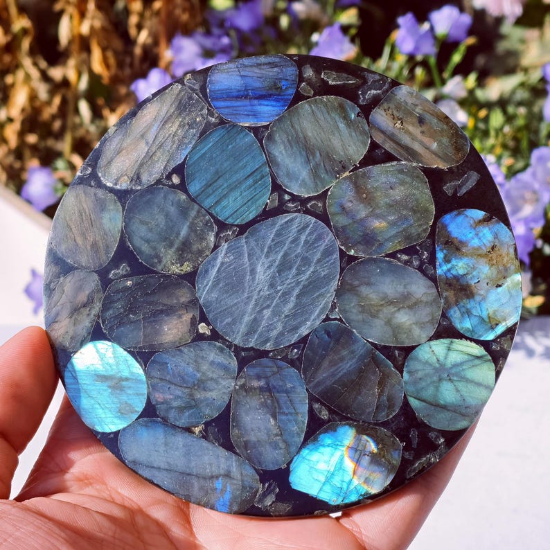 May include: A round coaster made of polished labradorite stones in shades of blue, teal, and gray, set in a dark matrix. The stones vary in size and shape, creating a mosaic-like pattern. The coaster is held in a hand.