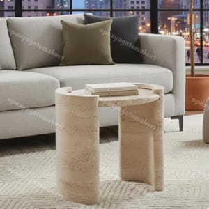 May include: A beige travertine side table with a rounded top and two curved legs. The table is approximately 46 cm in height and has a circular top. Two books are on the table. The table is in a living room setting.
