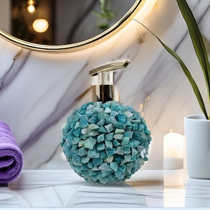 Puede incluir: Un dispensador de jabón decorativo con un cuerpo esférico cubierto de pequeñas piedras irregulares de color azul verdoso. El dispensador tiene una bomba plateada y está sobre una superficie blanca, con un fondo de mármol.