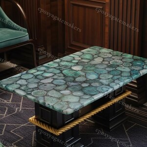 Op de afbeelding: Een rechthoekige salontafel met een moza&iuml;ekblad van groene en blauwe agaat schijfjes. De tafel heeft een zwart onderstel met gouden touwachtige accenten. De tafel is een uniek meubelstuk.