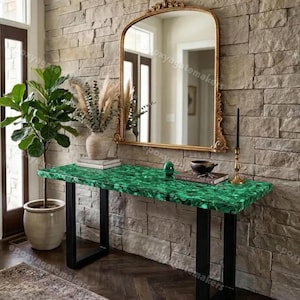 Op de afbeelding: Een console met een groene stenen blad en zwarte metalen poten. Boven de console hangt een goudkleurige spiegel. Op de console staan decoratieve items. De tafel is ongeveer 150 cm lang.
