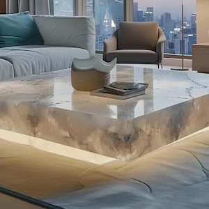 May include: A contemporary coffee table with a rectangular shape and a luminous, translucent surface. The table is constructed from a light-toned material, possibly stone or resin, and features integrated lighting. A sofa and armchair are visible.