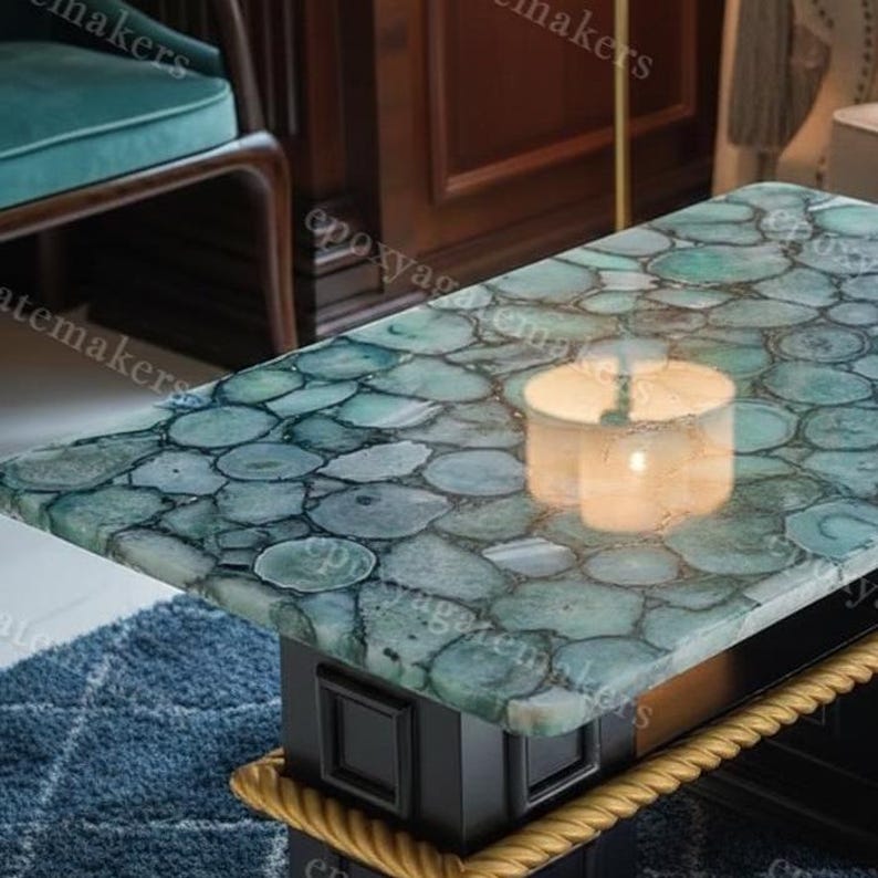 Op de afbeelding: Een rechthoekige salontafel met een blad van teal agaatsteen. De tafel heeft een zwarte basis met gouden touwrand. Een brandende kaars staat op de tafel en weerspiegelt in het stenen oppervlak. De tafel staat in een kamer met een blauw tapijt.