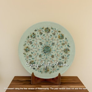 May include: A circular, pale blue decorative plate featuring an elaborate floral pattern in green, teal, and gold. The plate is displayed on a wooden stand. The text "Epoxy Agate Makers" is visible on the plate.
