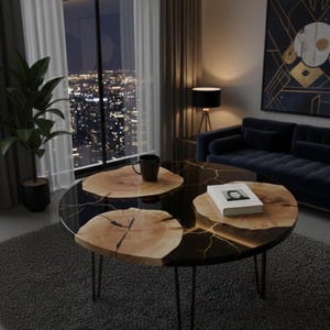 May include: Round coffee table with a black resin top and natural wood accents. The table features a unique design with gold-colored veining and slender metal legs. A book and a mug are placed on the table.