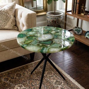 Könnte beinhalten: Ein runder Beistelltisch mit einer polierten Tischplatte aus grünem Achatstein. Die Tischplatte zeigt ein Mosaik aus grünen und weißen Achatscheiben. Der Tisch hat ein schwarzes Metallgestell im Dreibein-Stil. Der Tisch befindet sich in einem Wohnzimmer.