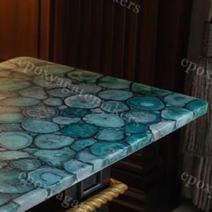 Op de afbeelding: Een close-up van een aanrechtblad met een design van blauwe en turquoise agaat. Het oppervlak is gemaakt van epoxyhars en heeft een patroon van cirkelvormige, steenachtige vormen. Het aanrechtblad heeft een glanzende afwerking en een donkere achtergrond.
