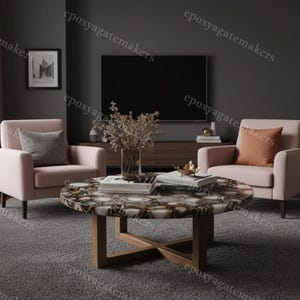 Puede incluir: Una mesa de centro redonda con una encimera de piedra de ágata única, sostenida por una base de madera. La mesa está rodeada por dos sillones rosas con cojines decorativos, un televisor y una imagen enmarcada. La habitación tiene un esquema de color neutro y moderno.