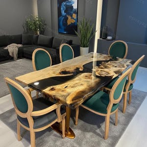 Può includere: Un tavolo da pranzo rettangolare con un design a bordo vivo, con un fiume di resina epossidica nera e venature del legno naturale. Il tavolo è circondato da sei sedie con sedili in velluto verde e telai in legno chiaro. Il tavolo è lungo circa 183 cm.