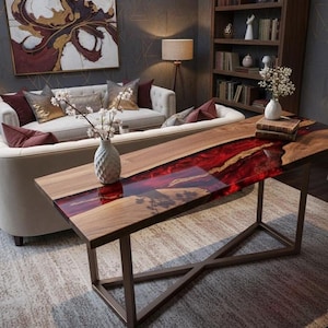 Puede incluir: Una mesa de centro rectangular de madera con un centro de resina roja vibrante. La mesa tiene un borde de madera marrón oscuro y se apoya sobre un marco de metal color bronce. La mesa está en un salón con un sofá blanco y objetos decorativos.