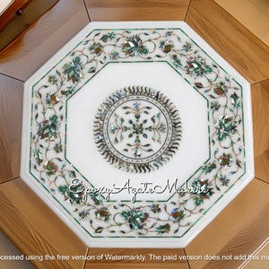 Puede incluir: Mesa octogonal de mármol blanco con intrincados incrustaciones florales en verde, marrón y acentos iridiscentes. El centro presenta un diseño circular. La mesa está sobre una base de madera.