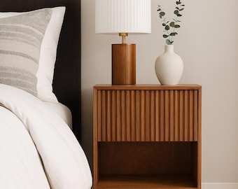 May include: A wooden nightstand with a ribbed front and a lower open shelf. It has gold-colored legs and a matching wooden lamp with a white pleated shade. A white vase with greenery sits on top.