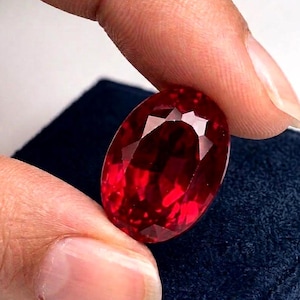 May include: A vibrant, oval-cut red gemstone with a faceted surface that reflects light. The stone displays a deep, saturated crimson hue and is held between fingers, showcasing its transparent quality and polished, symmetrical shape.