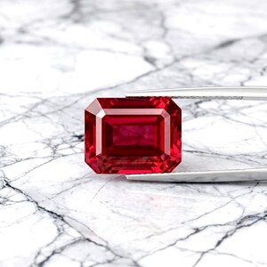May include: A vibrant, deep red gemstone cut in a rectangular emerald shape. The stone features precise, multi-faceted edges that reflect light, held securely by a pair of silver-toned metal tweezers against a white marble surface.