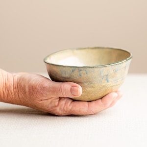 May include: A small, round, handmade ceramic bowl with a speckled beige and tan base. The upper portion of the bowl features a subtle, mottled blue glaze that blends into the natural clay tone, creating a textured, organic appearance.