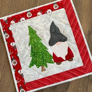 Może przedstawiać: Biało-szary kwadratowy kawałek tkaniny pikowanej z czerwoną i białą obwódką. W środku znajduje się zielone drzewko choinkowe i czerwono-biały krasnal w czarnym kapeluszu.