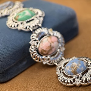 May include: A vintage silver-toned bracelet with oval-shaped, colorful stone settings. The bracelet features a series of ornate, filigree-style links, each holding a different colored stone: green, pink, and blue. The bracelet is displayed on a blue velvet surface.