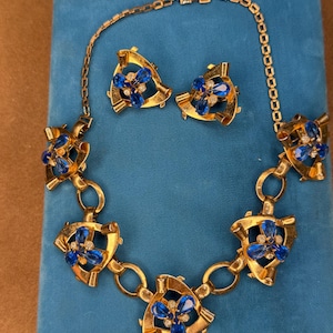 May include: A vintage jewelry set featuring a gold-toned necklace and matching earrings. The necklace and earrings showcase a design with blue gemstone accents and small clear crystals. The set is displayed on a blue velvet surface.