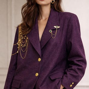 Puede incluir: Un blazer morado con botones dorados y un broche de abeja. El blazer tiene un corte entallado y un diseño clásico. El broche de abeja es dorado y verde, añadiendo un toque de elegancia. Un accesorio de cadena dorada para el hombro añade un toque único.