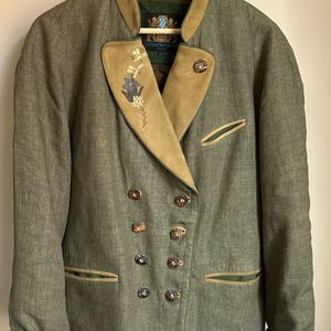 Puede incluir: Chaqueta verde de doble botonadura con cuello y puños de ante color beige. La chaqueta presenta botones decorativos y detalles florales bordados en la solapa. La chaqueta está colgada de una percha de madera.