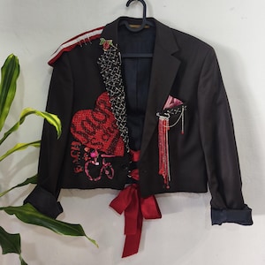 Puede incluir: Una chaqueta negra corta con un parche de corazón rojo con texto negro, una solapa cubierta de imperdibles plateados, un lazo rojo en la cintura y un broche de cadena plateada con cuentas rojas. El hombro presenta un adorno de rayas rojas y blancas.