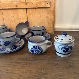 Puede incluir: Un juego de té de cerámica en miniatura con diseños florales azules. El juego incluye varias tazas pequeñas, platillos y un azucarero con tapa. Las tazas y los platillos tienen una base gris con patrones florales azules.
