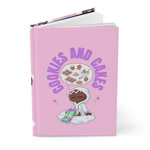 May include: A pink journal with the words "COOKIES AND CAKES" in a colorful font. The cover features an illustration of a person dreaming of baked goods. The person is wearing a white hoodie and sitting on a cloud.