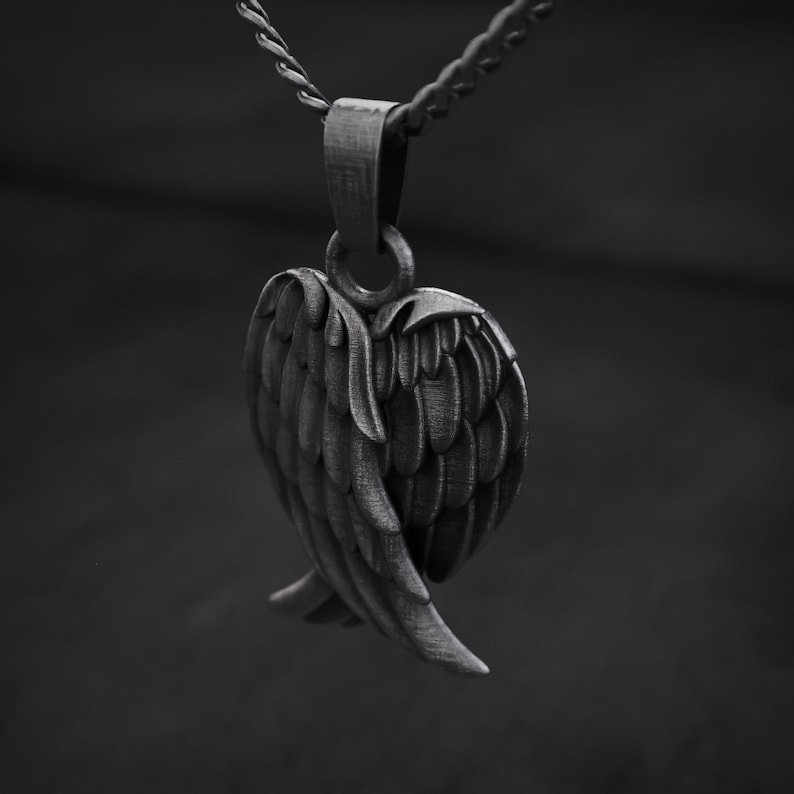 May include: A silver-toned pendant in the shape of a heart with detailed wing designs. The pendant is suspended from a matching chain, creating a monochromatic, elegant accessory. The wings are intricately detailed, adding depth and texture to the piece.