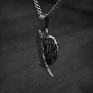 May include: A silver-colored pendant in the shape of a detailed angel wing, suspended from a matching chain. The wing features intricate feather detailing, creating a textured appearance. The background is a blurred, dark gray.