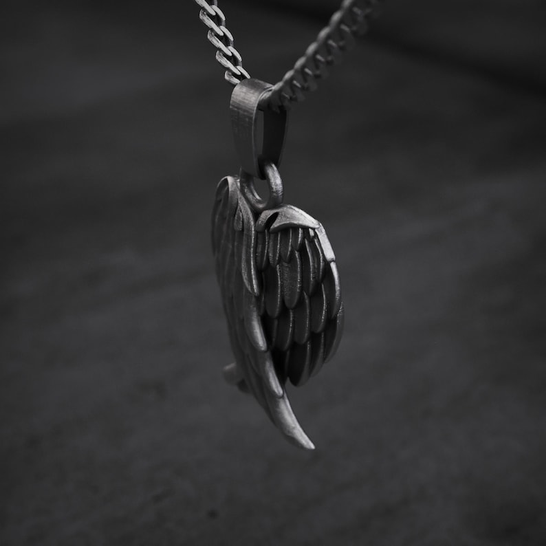 May include: A silver-colored pendant in the shape of a detailed angel wing, suspended from a matching chain. The wing features intricate feather detailing, creating a textured appearance. The background is a blurred, dark gray.