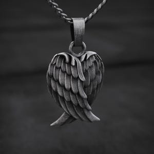 May include: A silver-toned pendant featuring a detailed wing design. The pendant is suspended from a matching chain. The wings are intricately sculpted, with each feather clearly defined. The piece is likely made of metal and is displayed against a dark background.