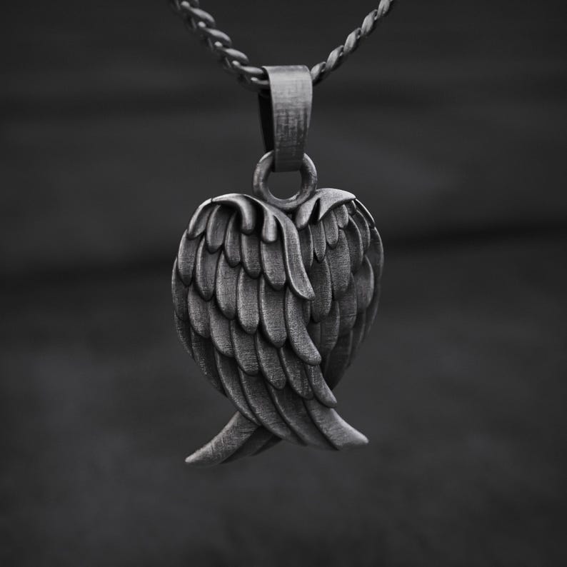 May include: A silver-toned pendant featuring a detailed wing design. The pendant is suspended from a matching chain. The wings are intricately sculpted, with each feather clearly defined. The piece is likely made of metal and is displayed against a dark background.