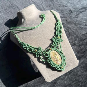 May include: A sage green macramé necklace featuring an oval-shaped, multi-coloured stone pendant at the centre. The intricate knotted cord design includes small, round, light-coloured beads integrated into the pattern, with a braided cord tie closure.
