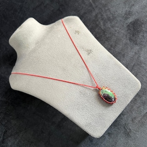 May include: A handmade necklace featuring a green and red stone pendant. The pendant is oval-shaped and wrapped in red thread, suspended from a matching red cord. The necklace is displayed on a gray mannequin.