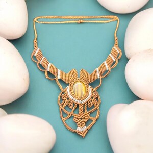 May include: A handcrafted macrame necklace with a large, oval, yellow and brown gemstone pendant. The necklace features intricate knotting in a golden-brown color, accented with white beads. The background is a teal color, surrounded by white stones.