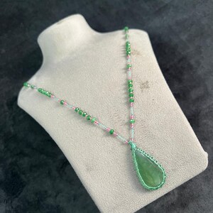 May include: A handcrafted necklace featuring a large, teardrop-shaped green pendant. The pendant is wire-wrapped and suspended from a delicate chain with green and pink beads. The necklace is displayed on a mannequin.