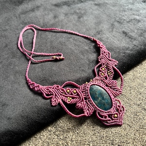 May include: A handmade macrame necklace in a deep magenta color. The necklace features an oval, teal-colored stone at the center, surrounded by intricate macrame knots and small gold-colored beads. The cord is adjustable.