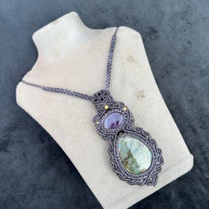 Puede incluir: Un collar de macramé hecho a mano con una gran piedra verde en forma de lágrima y una piedra púrpura ovalada más pequeña. Las piedras están engarzadas en un diseño de macramé gris oscuro con detalles amarillos, exhibido en un maniquí.