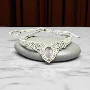 Peut inclure: Un collier ras du cou en macramé gris clair, orné d'une pierre précieuse ovale rose pâle au centre. Le collier est présenté sur une pierre grise. Le design en macramé est complexe et détaillé.