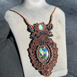 Op de afbeelding: Een handgemaakte macramé ketting met een grote, ovale labradoriet edelsteen met iriserende kleurschitteringen. De hanger is ingewikkeld geweven met bruin en zwart koord, geaccentueerd door een kleine turkoois steen. De ketting wordt getoond op een grijze buste.