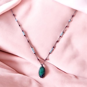 Może przedstawiać: Ręcznie robiony naszyjnik z ciemnozieloną owalną zawieszką i ciemnofioletową obwódką. Naszyjnik ma czarny sznurek z naprzemiennymi małymi czerwonymi i jasnoniebieskimi koralikami. Tło to miękka różowa tkanina.