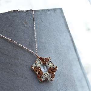 May include: A handmade macrame necklace with a star-shaped pendant. The pendant features a clear, round center surrounded by brown, cream, and gold-colored macrame detailing. The necklace hangs on a delicate, light-colored chain.