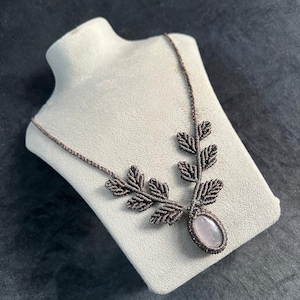Puede incluir: Un collar de macramé hecho a mano con un diseño de hojas y una piedra central ovalada de cuarzo rosa. El collar se exhibe sobre un busto blanco contra un fondo oscuro. El macramé es de color marrón oscuro.
