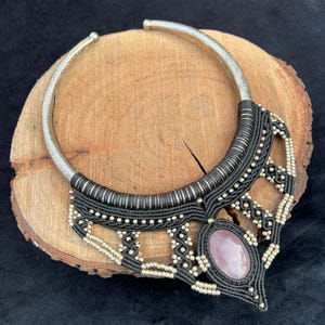 Peut inclure: Collier ras du cou argenté avec un motif macramé noir et une grosse pierre rose ovale. Le collier est présenté sur une surface en bois.