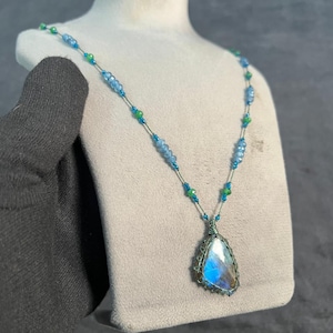Peut inclure: Un collier artisanal avec un pendentif en forme de goutte d'eau, orné d'une pierre irisée bleue et blanche dans une monture en fil de fer. La chaîne délicate est ornée de perles bleues et vertes, présentée sur un mannequin.